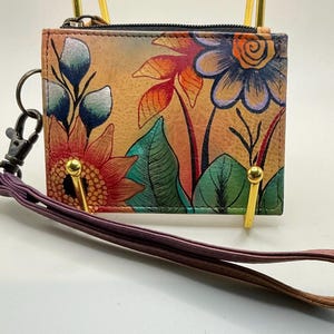 Puede incluir: Una cartera pequeña y rectangular con un diseño floral que presenta girasoles y otras flores coloridas. La cartera tiene una cremallera negra, herrajes dorados y una correa de muñeca morada. El fondo es de un tono cálido y terroso.
