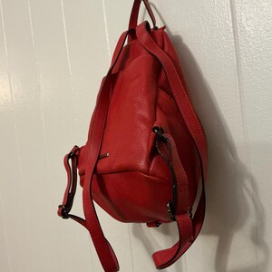 May include: A bright red leather backpack with a textured surface, featuring adjustable shoulder straps, a top carry handle, and dark metal hardware accents. The bag has a rounded base and a vertical orientation, showing the back panel and straps.