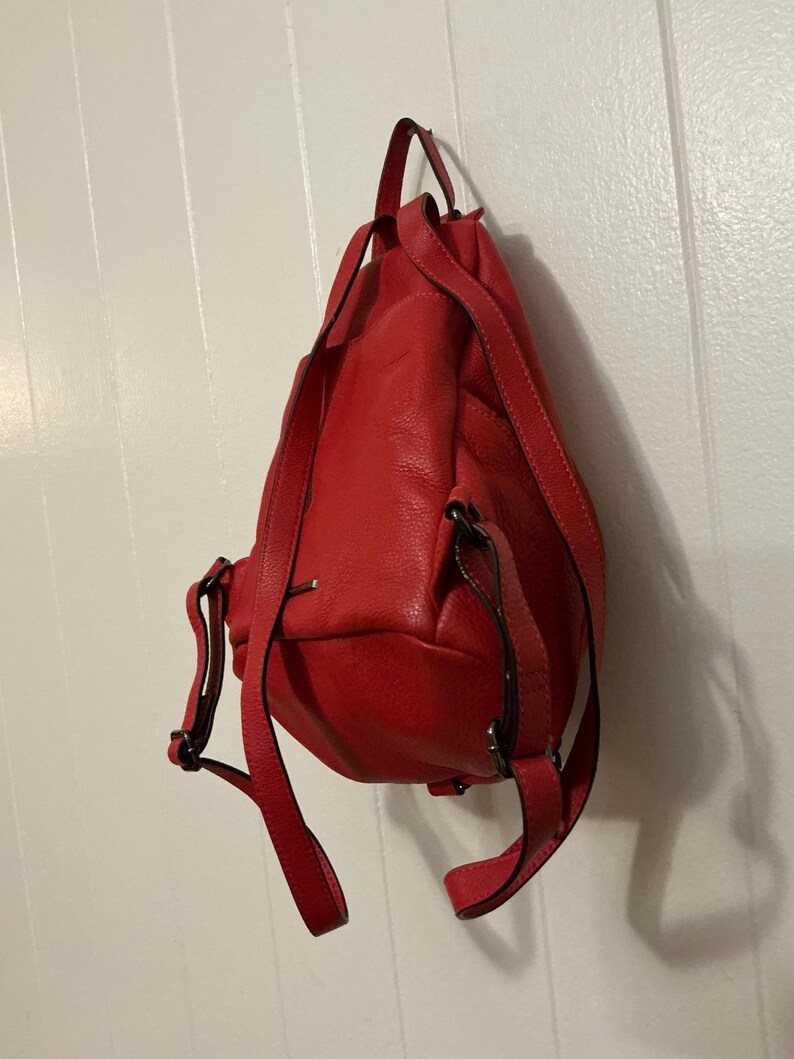 May include: A bright red leather backpack with a textured surface, featuring adjustable shoulder straps, a top carry handle, and dark metal hardware accents. The bag has a rounded base and a vertical orientation, showing the back panel and straps.