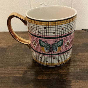 May include: A ceramic mug with a gold handle and rim. The mug features a pink background with a mosaic tile pattern in gold, white, and black. A butterfly design is centered on the mug.
