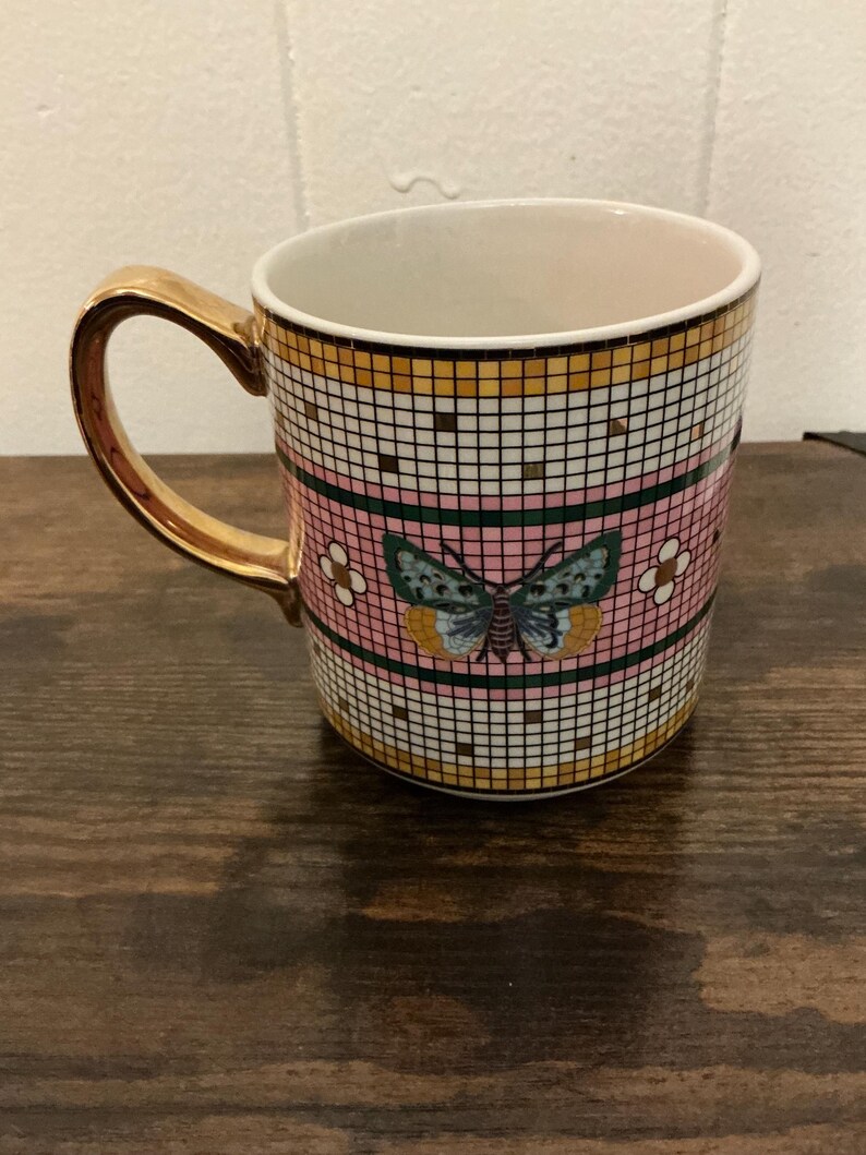 May include: A ceramic mug with a gold handle and rim. The mug features a pink background with a mosaic tile pattern in gold, white, and black. A butterfly design is centered on the mug.