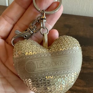 May include: A heart-shaped keychain covered in shimmering gold sequins. A horizontal beige fabric band runs across the center, featuring the embossed text LeSportsac. The heart is attached to a silver-toned metal split ring and a lobster clasp.