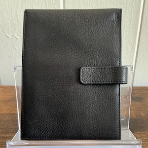 May include: A black leather journal with a textured surface and a rectangular shape. The journal has a strap closure on the right side. The journal is displayed on a clear acrylic stand.
