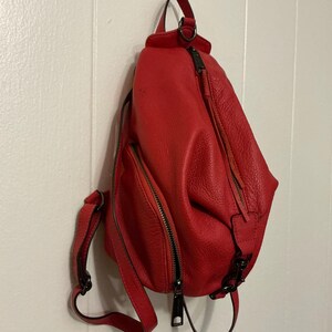 May include: A bright red, textured leather sling backpack featuring a teardrop shape, a vertical zippered front pocket, a main zippered compartment, and adjustable shoulder straps with dark metal hardware and matching red leather zipper pulls.
