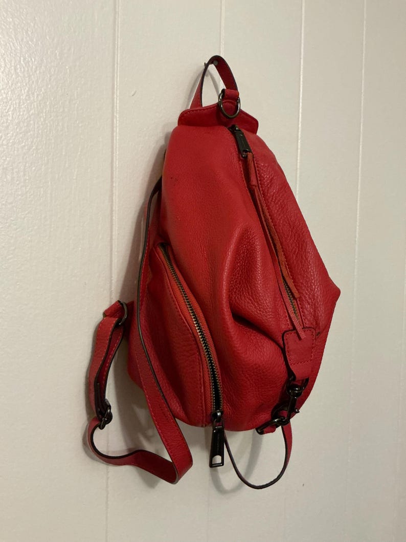 May include: A bright red, textured leather sling backpack featuring a teardrop shape, a vertical zippered front pocket, a main zippered compartment, and adjustable shoulder straps with dark metal hardware and matching red leather zipper pulls.