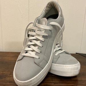 May include: A pair of light gray sneakers with white laces and soles. The shoes have a low-profile design with a rounded toe and a padded collar. The sneakers are made of a suede-like material and are suitable for casual wear.