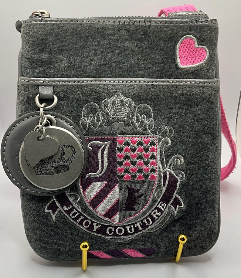 May include: Gray crossbody bag with a pink heart patch and a pink strap. The bag features a decorative crest with the words "Juicy Couture" embroidered in white. A silver charm with a heart and crown hangs from the bag.
