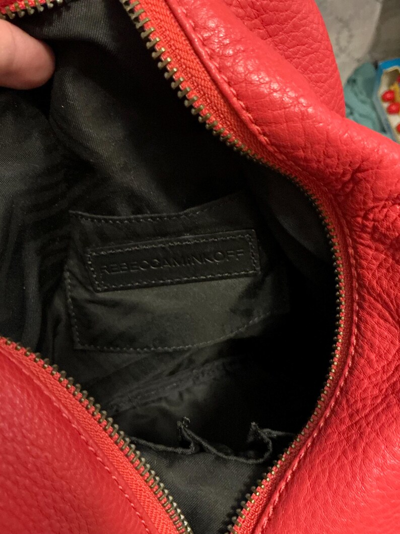 May include: A bright red textured handbag with a gold-toned zipper closure. The interior is lined with black fabric and features a rectangular black leather patch embossed with the text REBECCAMINKOFF, along with small internal pockets.