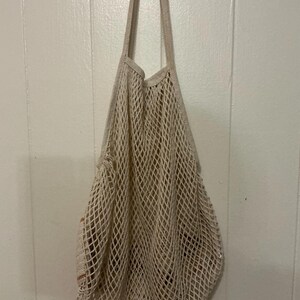 May include: A beige cotton string market bag featuring an open-weave mesh design with a diamond pattern. The bag has two integrated shoulder straps and a flexible, expandable body structure that hangs vertically when empty or filled with items.