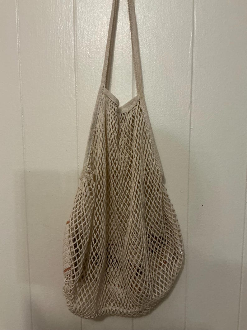 May include: A beige cotton string market bag featuring an open-weave mesh design with a diamond pattern. The bag has two integrated shoulder straps and a flexible, expandable body structure that hangs vertically when empty or filled with items.