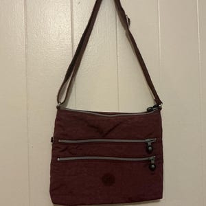 May include: A burgundy crossbody bag with a brown shoulder strap. The bag has three zippered compartments and a small circular logo on the front. The bag is hanging against a white wall.