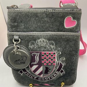 May include: Gray velour crossbody bag with a pink strap and heart accent. The bag features an embroidered crest with the words "Juicy Couture" and a silver-tone charm with a crown design.