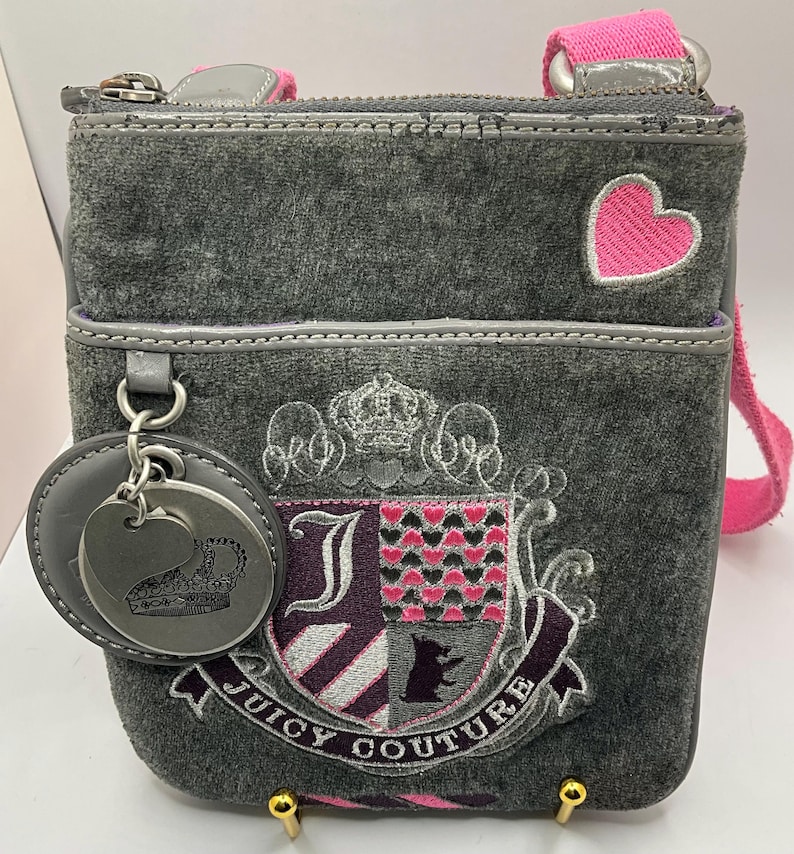 May include: Gray velour crossbody bag with a pink strap and heart accent. The bag features an embroidered crest with the words "Juicy Couture" and a silver-tone charm with a crown design.