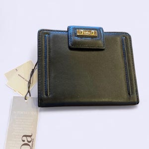 May include: Black leather wallet with a gold-tone clasp and a small gold-tone logo. The wallet has a rectangular shape with a flap closure. A tag with the words "Genuine Leather" is attached.
