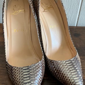 Puede incluir: Un par de zapatos de tacón alto con estampado de piel de serpiente en tonos marrón y beige. Los zapatos tienen punta afilada y tacones de aguja. Las plantillas son de color beige claro con letras doradas.