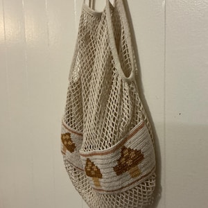 May include: A cream-colored crochet net bag featuring a solid band at the base decorated with brown and tan mushroom motifs. The bag has two long shoulder straps and a wide, open-weave mesh body constructed from durable, lightweight yarn.