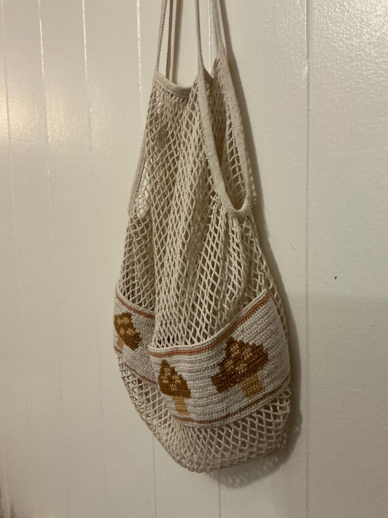 May include: A cream-colored crochet net bag featuring a solid band at the base decorated with brown and tan mushroom motifs. The bag has two long shoulder straps and a wide, open-weave mesh body constructed from durable, lightweight yarn.