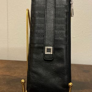 May include: A black leather card holder wallet featuring multiple horizontal card slots, a vertical strap with a silver square metal emblem, and a side zipper closure. The wallet has a slim, rectangular design with visible stitching along the edges.