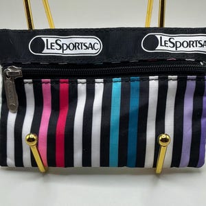 Puede incluir: Un estuche rectangular LeSportsac con cremallera, con la parte superior negra y un patrón de rayas verticales en negro, blanco, rosa, rojo, turquesa y morado. El logotipo de LeSportsac está impreso en la parte superior negra. El estuche está sujeto por dos soportes metálicos dorados.