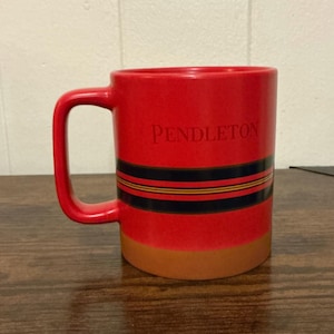 Puede incluir: Una taza de cerámica de color rojo brillante con la palabra PENDLETON impresa en fuente serif. La taza tiene una banda horizontal de rayas negras y finas rayas amarillas en el centro, una base de color tostado y un asa de color rojo.
