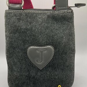 May include: Gray crossbody bag with a pink strap and a heart-shaped patch featuring the letter "J". The bag has a zippered top and gold-colored feet. The bag is made of a soft, velvety material.
