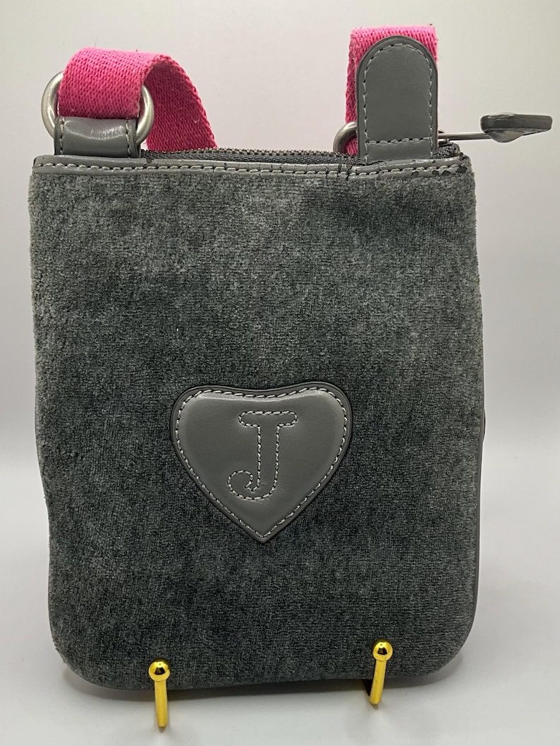May include: Gray crossbody bag with a pink strap and a heart-shaped patch featuring the letter "J". The bag has a zippered top and gold-colored feet. The bag is made of a soft, velvety material.