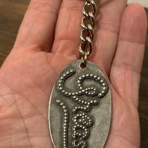 Può includere: Portachiavi color argento con un ciondolo ovale. Il ciondolo presenta la parola "peace" in un carattere stilizzato e puntinato. Il portachiavi include una catena e un anello diviso per attaccare le chiavi.