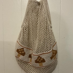 May include: A cream-colored crochet net bag featuring a solid band around the lower section decorated with a repeating pattern of brown and tan mushrooms. The bag has two integrated handles and a flexible, open-weave mesh construction.