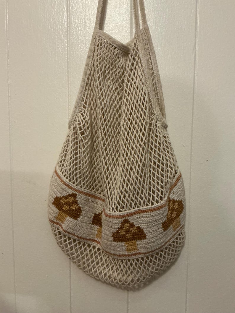 May include: A cream-colored crochet net bag featuring a solid band around the lower section decorated with a repeating pattern of brown and tan mushrooms. The bag has two integrated handles and a flexible, open-weave mesh construction.
