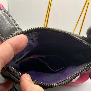 May include: A gray leather crossbody bag with a pink strap. The bag has a zippered main compartment and a small pocket inside. A silver heart-shaped charm hangs from the zipper. The interior lining is purple with a paisley pattern.