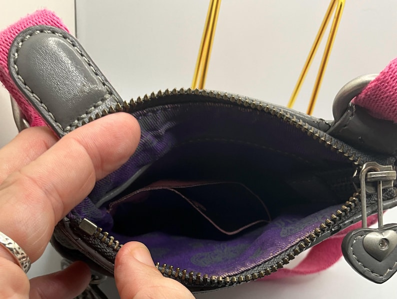 May include: A gray leather crossbody bag with a pink strap. The bag has a zippered main compartment and a small pocket inside. A silver heart-shaped charm hangs from the zipper. The interior lining is purple with a paisley pattern.
