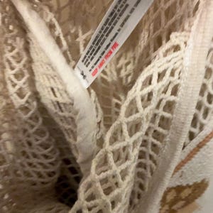 May include: A cream-colored mesh tote bag featuring a diamond-patterned knit construction. A white fabric care label is attached to the interior seam, displaying printed text including the warning KEEP AWAY FROM FIRE in red capital letters.