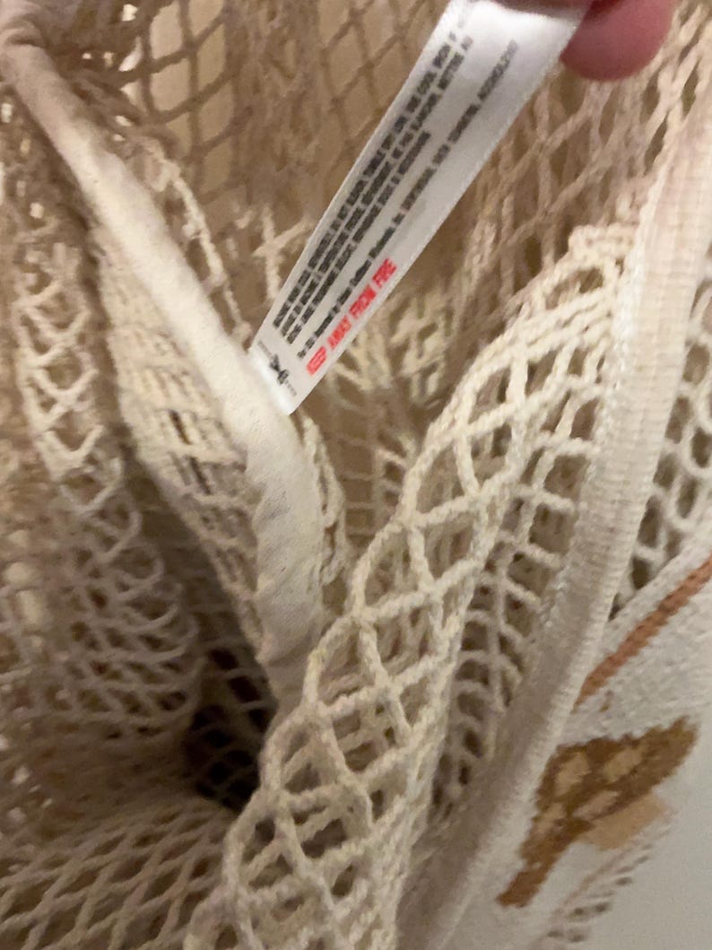 May include: A cream-colored mesh tote bag featuring a diamond-patterned knit construction. A white fabric care label is attached to the interior seam, displaying printed text including the warning KEEP AWAY FROM FIRE in red capital letters.