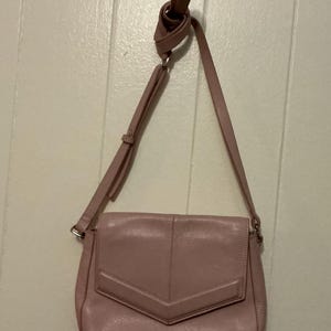May include: A dusty rose-colored crossbody bag with a flap closure and adjustable strap. The bag features a geometric design on the front flap and silver-toned hardware. The strap is looped at the top.