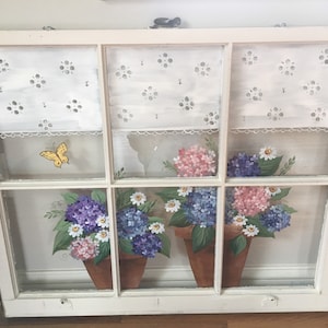 May include: A repurposed window frame with a painted floral design. The artwork features pots of hydrangeas in shades of pink, purple, and blue, with white daisies. A yellow butterfly and white lace curtains complete the scene.