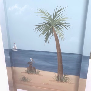 May include: A hand-painted wooden cabinet door depicting a beach scene. The scene includes a blue sky with a white cloud, a palm tree, a blue ocean, a sailboat, a sandy beach, and a pelican perched on a wooden post.