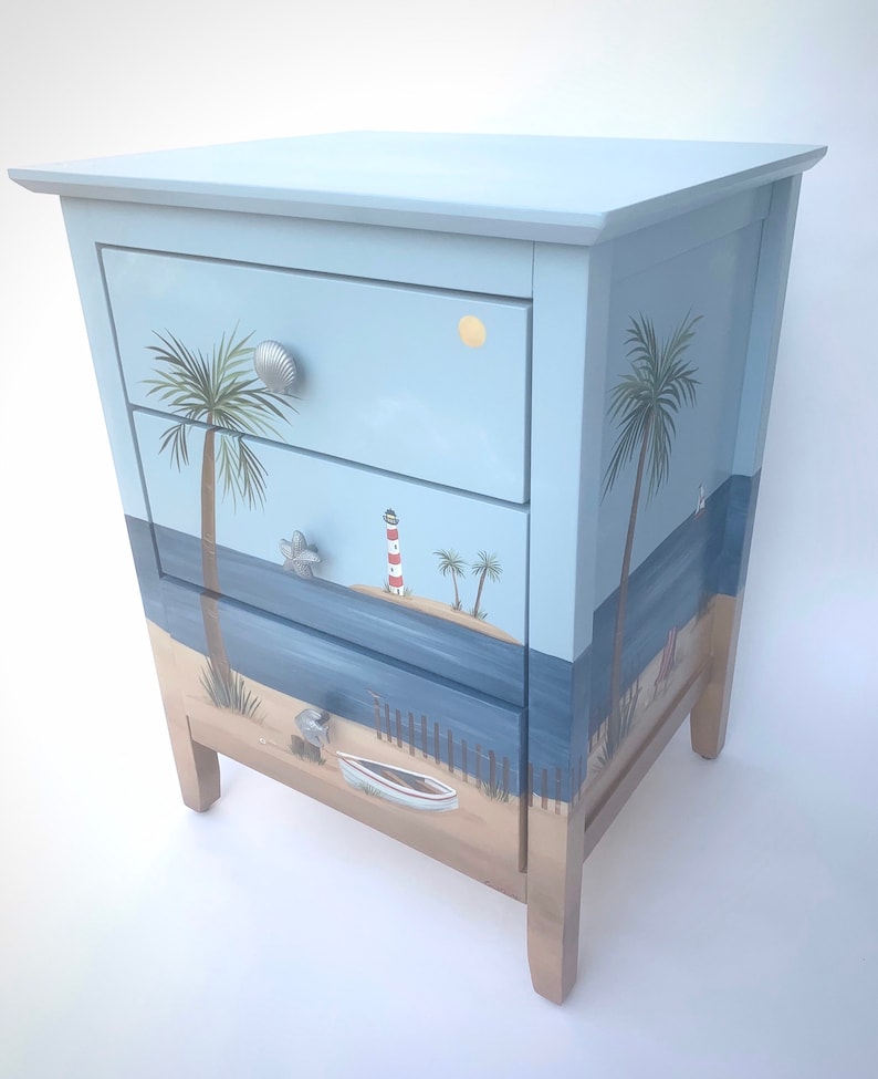 May include: A light blue wooden chest of drawers with three drawers. The drawers are decorated with a beach scene, including palm trees, a lighthouse, a boat, and a sandy beach.
