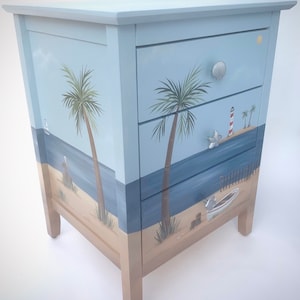 May include: A light blue wooden chest of drawers with three drawers. Each drawer features a painted beach scene with palm trees, a lighthouse, and a boat. The dresser has silver drawer pulls.