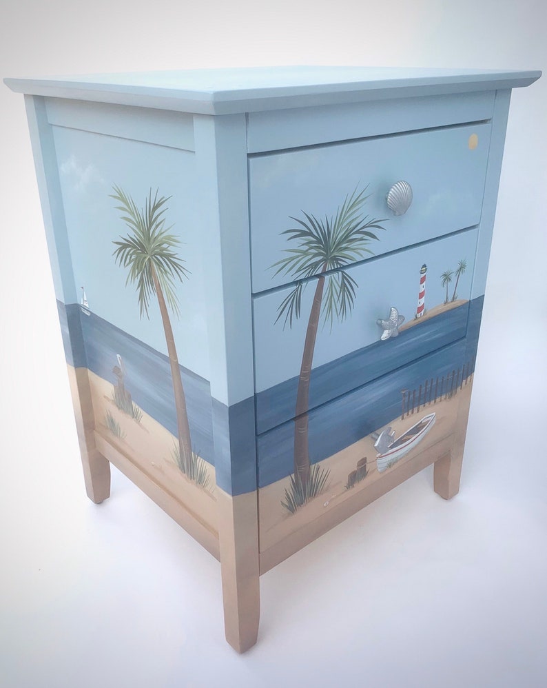 May include: A light blue wooden chest of drawers with three drawers. Each drawer features a painted beach scene with palm trees, a lighthouse, and a boat. The dresser has silver drawer pulls.
