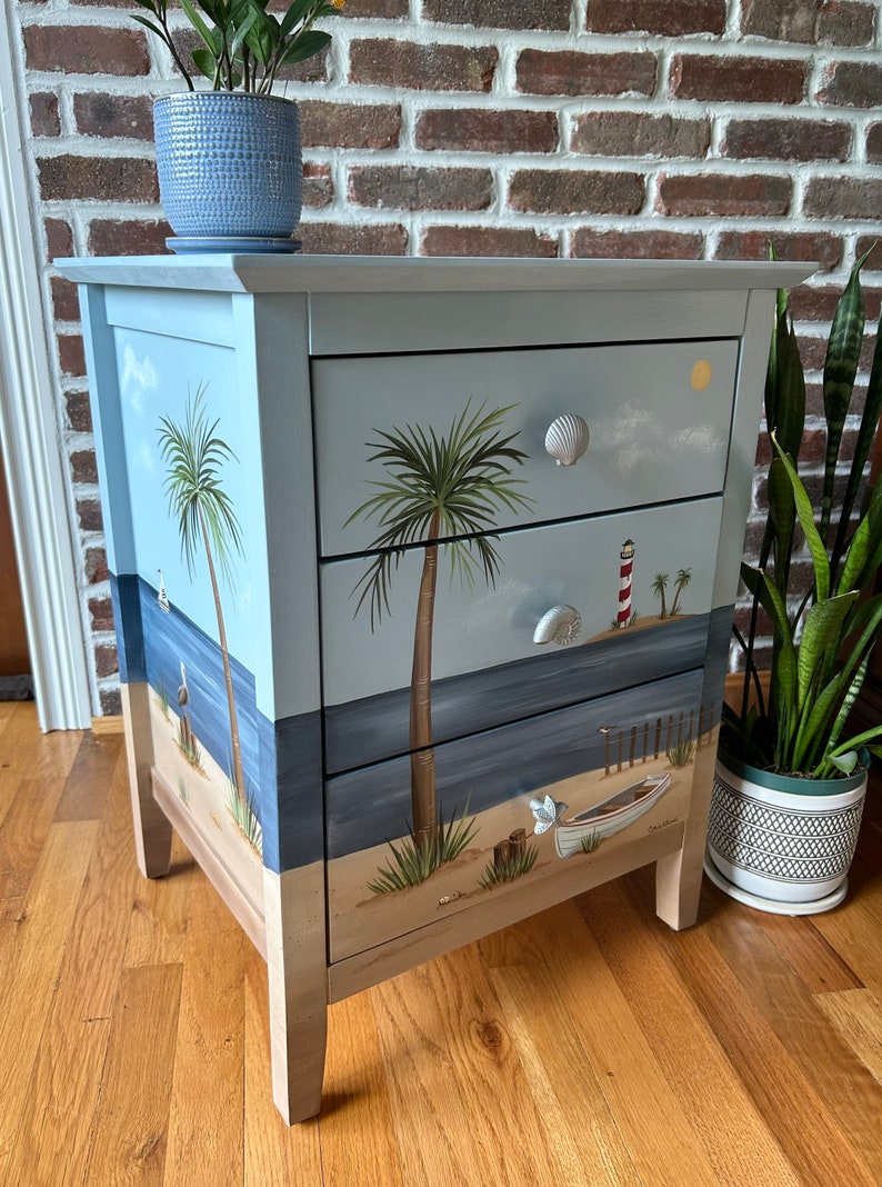May include: A three-drawer chest of drawers painted with a beach scene. The drawers feature a blue sky, palm trees, a lighthouse, a boat, and a sandy beach. The dresser has white seashell-shaped drawer pulls.