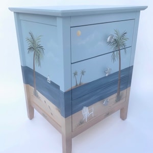 May include: A three-drawer chest of drawers painted with a beach scene. The drawers feature painted palm trees, a sailboat, a beach chair, and seashells. The chest is painted in shades of blue, green, and brown.