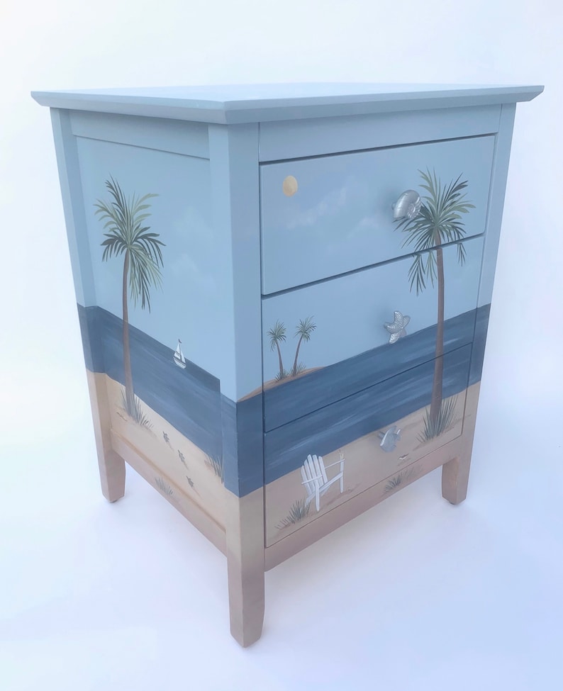 May include: A three-drawer chest of drawers painted with a beach scene. The drawers feature painted palm trees, a sailboat, a beach chair, and seashells. The chest is painted in shades of blue, green, and brown.