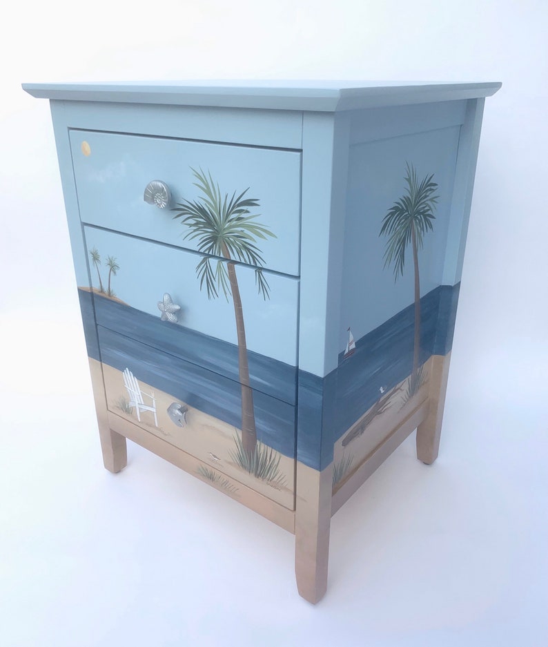 May include: A light blue wooden chest of drawers with three drawers. The drawers are painted with a beach scene featuring palm trees, a sailboat, and a beach chair. The chest of drawers has a light brown base.
