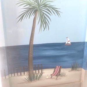 May include: A hand-painted wooden chair featuring a beach scene. The scene depicts a palm tree, a sailboat, a beach chair, and a drink on the sand.