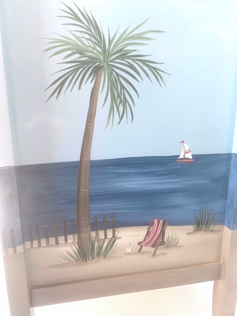 May include: A hand-painted wooden chair featuring a beach scene. The scene depicts a palm tree, a sailboat, a beach chair, and a drink on the sand.