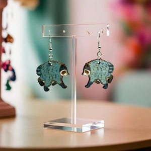 May include: A pair of dangle earrings featuring a blue raccoon illustration with dark grey markings and a white face. The earrings are suspended from silver-toned metal hooks and are displayed on a clear acrylic T-shaped jewelry stand.