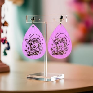 Könnte beinhalten: Ein Paar rosa tropfenförmige Ohrringe aus Holz mit einem lasergravierten Bild von Dolly Parton, die einen Cowboyhut trägt, und dem Wort "Dolly" darunter.