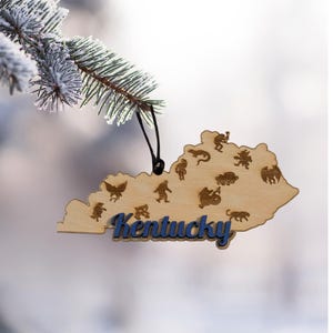 May include: A wooden Christmas ornament shaped like the state of Kentucky, with blue lettering that spells "Kentucky". The ornament is decorated with various small cut-out figures and is hanging from a black string.