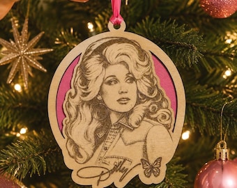 May include: A circular wooden ornament featuring a laser-engraved portrait of a woman with voluminous hair, wearing a collared shirt. The design includes a signature and a small butterfly graphic, set against a vibrant pink background with a pink ribbon.