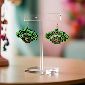Peut inclure: Une paire de boucles d'oreilles Cabbage Patch Kids sur un présentoir transparent. Les boucles d'oreilles sont vertes et noires, avec le logo Cabbage Patch Kids et un motif de chou. Les boucles sont suspendues à des crochets dorés.